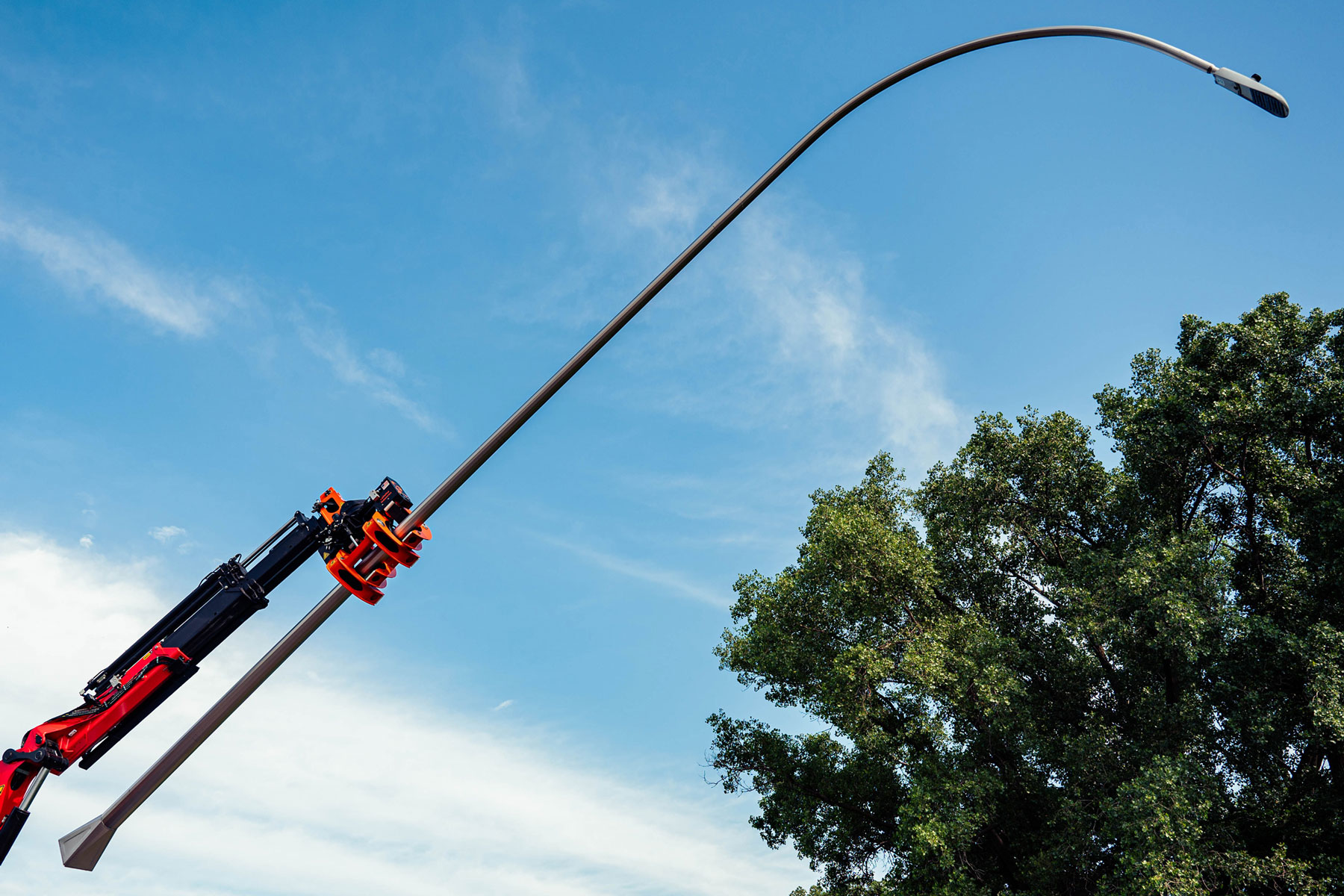 The PM1000™ pole grapple is pictured attached to a crane truck. It is gripping a large, curved light pole during placement for installing on an anchor. There is a large tree in the lower right corner.