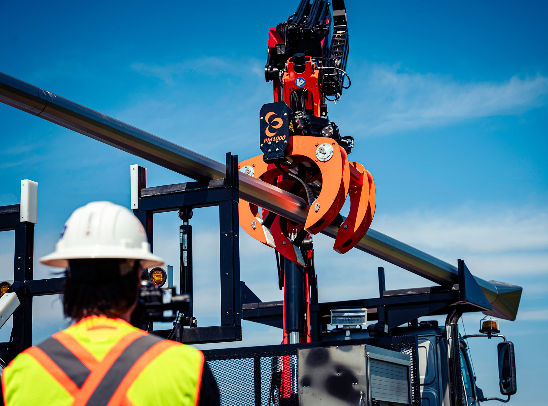The PM1000™ pole grapple by LaValley Industries is about to grab a pole from a rack. The grapple is attached to a crane truck, and the rack is on the back of the truck. There is an operator in the foreground with their back turned to the camera.